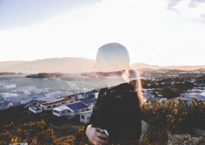 A double exposure of a young woman's side profile portrait and a landscape with hills and houses at sunset